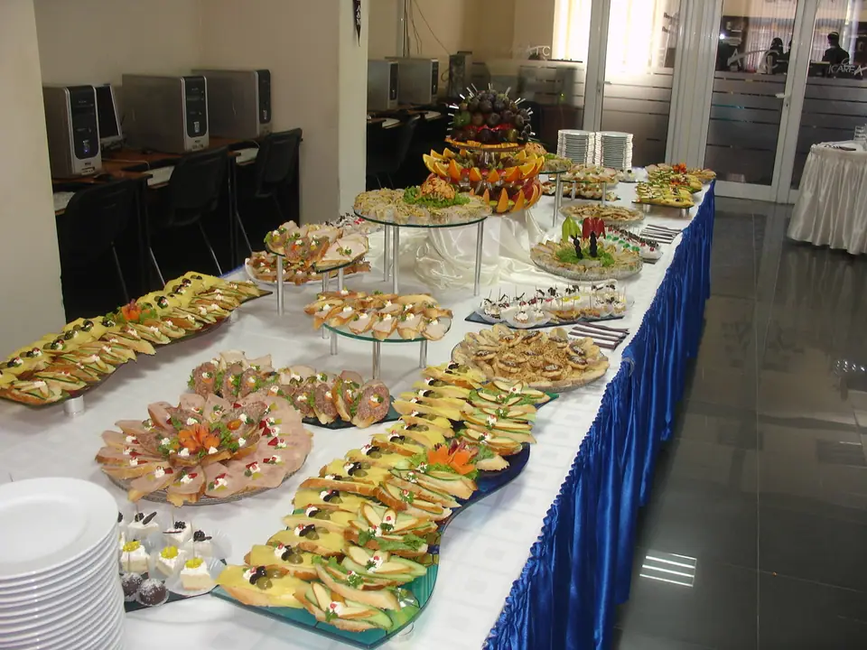 Mesa larga de buffet con mantel blanco y bandejas de bocadillos, sándwiches y fiambres