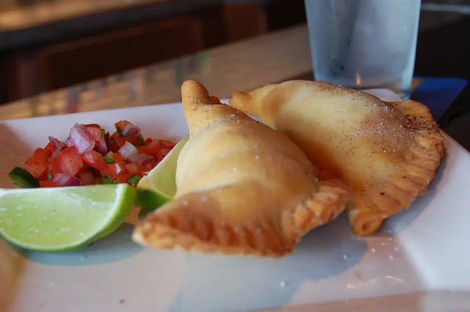 Dos empanadas doradas con repulgue en un plato, junto a salsa de tomate y lima