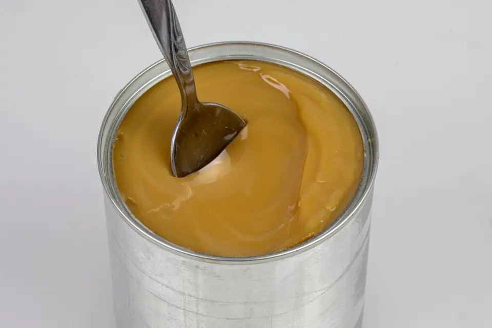Lata abierta de leche condensada caramelizada con una cuchara adentro, sobre fondo blanco