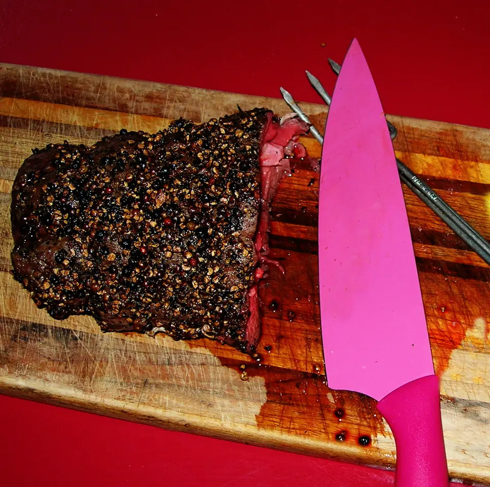 Lomo asado entero con costra de pimienta, cortado en un extremo, sobre una tabla de madera junto a un cuchillo