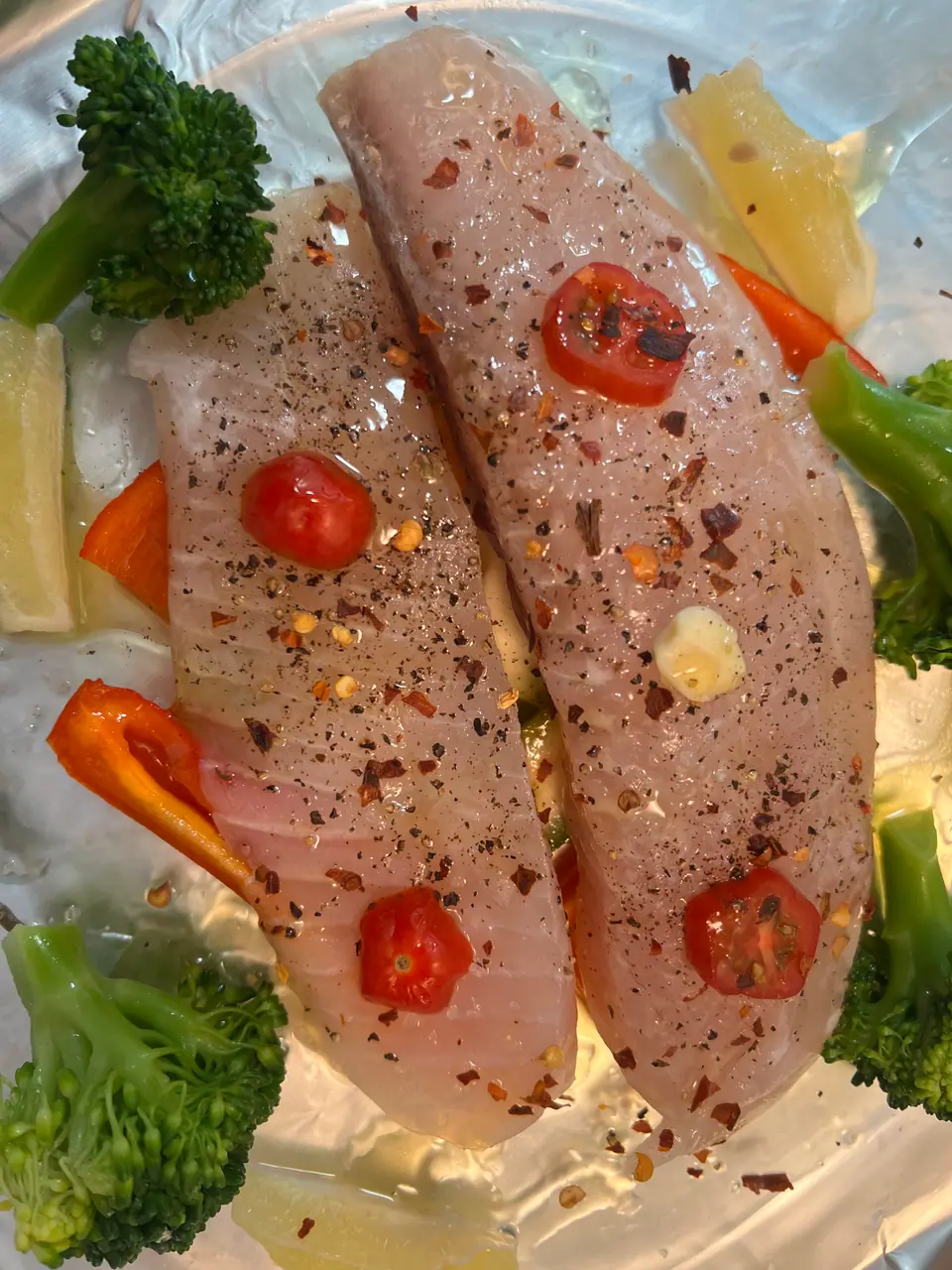 Dos filetes de pescado blanco crudos condimentados, con tomates cherry y brócoli, listos para hornear