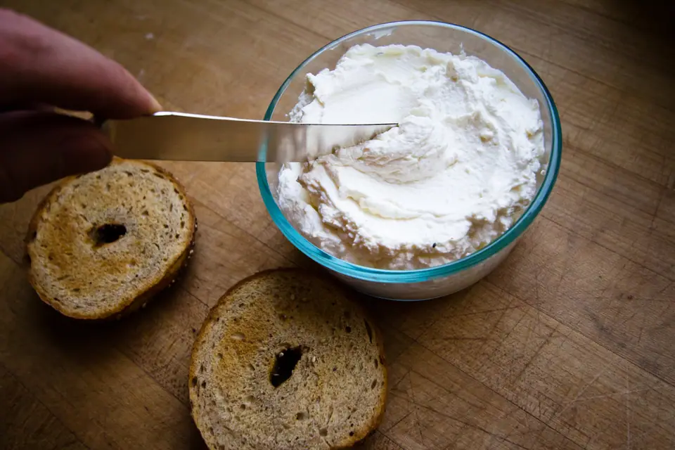 Bol de vidrio con queso crema y un cuchillo untándolo, junto a dos mitades de bagel tostado